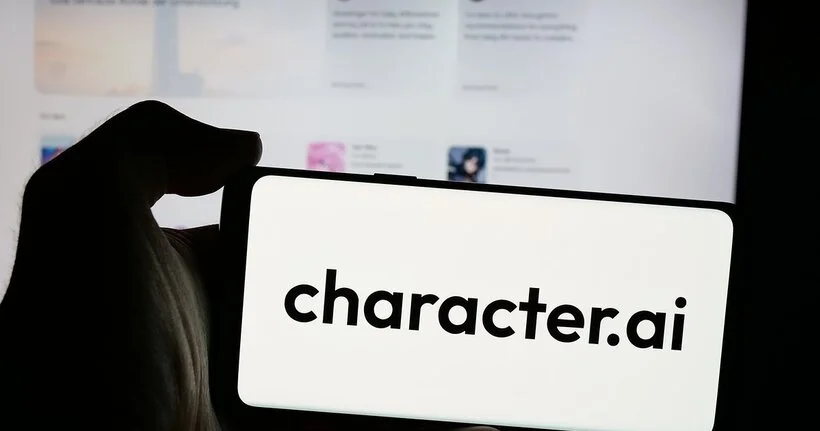 A hand holds a smartphone that reads character.ai in front of a computer screen.